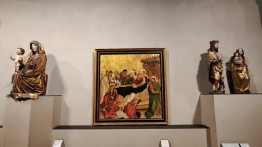 Rönesans dönemi dini resimler ve heykeller sergilenen, incildeki detaylı sahneler ve hafif ışıklandırma altında sergilenen süslü altın arka planlar sergilenen bir müze galerisi.
