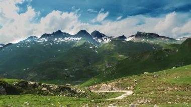 Yaz boyunca dağlık bir bölgenin görkemli manzarası, açık mavi gökyüzü altında karla kaplı dağlarla zıt yemyeşil çayırları gösteriyor.