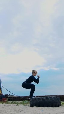 Odaklanmış bir kadın dramatik bir gökyüzü altında büyük bir tekerleği devirmeye hazırlanan dinamik bir egzersize girişir. Yoğunlaşmış çabası, açık hava egzersizleri sırasında azim ve gücü yansıtıyor..