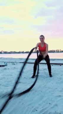 Kararlı bir kadın kumlu bir plajda savaş halatlarıyla egzersiz yapıyor, çarpıcı bir günbatımına karşı. Onun odaklanmış ifadesi ve dinamik hareketleri güç ve adanmışlığı iletir..