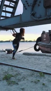 Kararlı bir kadın sahildeki çelik bir yapının altında süspansiyon kayışları kullanarak antrenman yapıyor. Akşam gökyüzü ve sakin su dingin bir zemin oluşturur, odak ve güç verir..