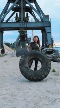 Bir kadın, kumlu bir plajda lastik egzersizi yaparken güç ve odaklanma gösteriyor. Endüstriyel vinç arka planı sahnenin dinamik enerjisini arttırmak için eşsiz bir kontrast ekliyor..