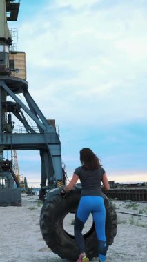 Bu dinamik sahnede, odaklanmış genç bir kadın endüstriyel bir rıhtımda büyük lastiklerle çalışıyor. Akşamın erken saatlerinde gökyüzü ve sakin su ilham verici, esnek bir hava yaratır..