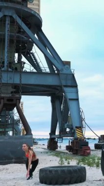 Kırmızı bluzlu ve siyah taytlı atletik bir kadın büyük bir traktör tekerleği kullanarak farklı güç egzersizleri yapar, kaslarını çalıştırır. Arkasında kargo vinci, kargo limanı, şafak