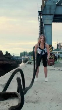 Odaklanmış bir kadın, rıhtımda iplerle savaşarak güçlü bir şekilde egzersiz yapar. Endüstriyel yapı, onun enerjisi ve kararlılığı, gücü ve dayanıklılığıyla zıtlaşıyor..