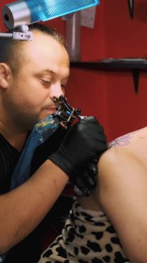 Dövme salonunda bir uzman, kadının sırtına çiçek süsü dövmesi yaptırıyor. Bir adam özel eldivenlerle, özel ekipmanlarla çalışır. Çizim siyah boyayla yapılır..