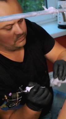 Dövme salonunda bir uzman, kadının sırtına çiçek süsü dövmesi yaptırıyor. Bir adam özel eldivenlerle, özel ekipmanlarla çalışır. Çizim siyah boyayla yapılır..