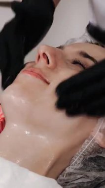Güzellik terapisi. Kozmetik cilt bakımı. Siyah tıp eldivenli güzellik uzmanı kozmetik cilt bakımı yapıyor, temizleyici köpük ya da kadın yüzüne emülsiyon uyguluyor. Yakın plan. Yaşlanma karşıtı prosedür. Yüz tazeleme.