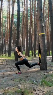 Atletik, eğitmen, egzersiz yapıyor, fitness sistemiyle egzersiz yapıyor, süspansiyon kayışları takıyor. Çam ormanlarında, yazın, güneş ışınlarında.