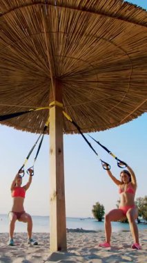 İki atletik, mayolu seksi genç bayan, eğitmenler, fitness sistemi ile egzersiz yapıyorlar, süspansiyon kayışları. Kumsalda, yazın, gün batımında, güneş ışınlarında.