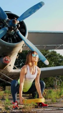 Atletik, güneş gözlüklü, taytlı genç bir kadın, eski bir uçağın arka planında ağır ağır bir plakayla egzersiz yapıyor.
