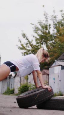seksi atletik genç sarışın kadın şort, eski terk edilmiş garajlar yakınındaki yaz aylarında lastikler, şınav, yardımıyla çeşitli güç egzersizleri gerçekleştirir