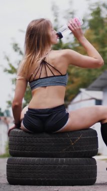 Güzel seksi atletik genç sarışın bir kadın üst ve crossfit egzersiz yaptıktan sonra lastik ve içme suyu bir şişe oturan şort. Yaz aylarında yakınındaki eski terk edilmiş garajlar.