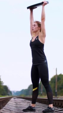 Siyah taytlı bir kadın ağır bir plakayla egzersiz yapıyor demiryolunda, raylarda, uyku tulumlarında, yazın. Yavaş çekim