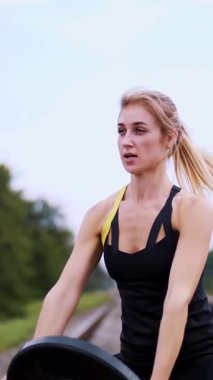 Siyah taytlı bir kadın ağır bir plakayla egzersiz yapıyor demiryolunda, raylarda, uyku tulumlarında, yazın. Yavaş çekim