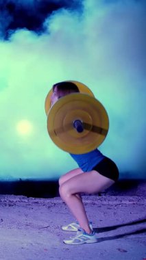 Atletik kız, halteri kaldır, halterle mekik çek. Geceleyin, projektörlerin ışığında, hafif dumanlarda, siste, terk edilmiş eski bir hangarda, bina.