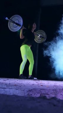 Atletik kız, halteri kaldır, halterle mekik çek. Geceleyin, projektörlerin ışığında, hafif dumanlarda, siste, terk edilmiş eski bir hangarda, bina.