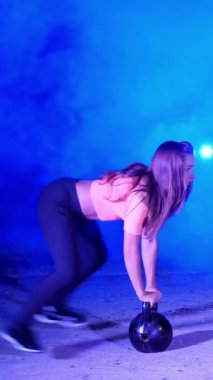 Ağırlıkları ile çeşitli güç egzersizleri yapıyor atletik, güzel genç bir kadın. Geceleri, projektörler, hangarda bir eski terk edilmiş, Bina hafif duman, sis ışığında.