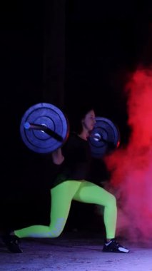 Atletik, genç kadının bir halter, lunges, ile çeşitli güç egzersizleri yapması ağız kavgası. Duman, sis, çok renkli projektörler ışığında gece içinde binayı terk edilmiş bir