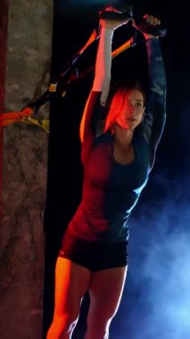 Atletik kadın spor yapıyor spor sistemi, süspansiyon kayışları. Geceleri, hafif dumanlarda, siste, çok renkli projektörlerde, terk edilmiş binada.