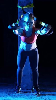 Atletik kadın spor yapıyor spor sistemi, süspansiyon kayışları. Geceleri, hafif dumanlarda, siste, çok renkli projektörlerde, terk edilmiş binada.