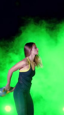 Ağırlıkları ile çeşitli güç egzersizleri yapıyor atletik, güzel genç bir kadın. Geceleri hafif duman, sis, çok renkli projektörler ışığında içinde binayı terk edilmiş