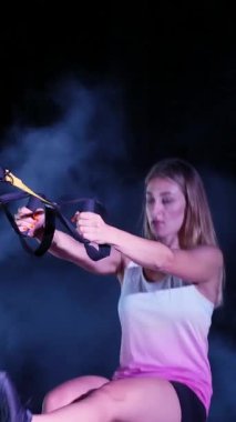 Atletik kadın spor yapıyor spor sistemi, süspansiyon kayışları. Geceleri, hafif dumanlarda, siste, çok renkli projektörlerde, terk edilmiş binada.