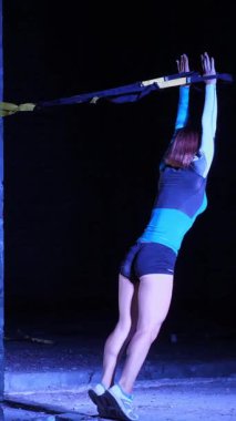 Atletik kadın spor yapıyor spor sistemi, süspansiyon kayışları. Geceleri, hafif dumanlarda, siste, çok renkli projektörlerde, terk edilmiş binada.