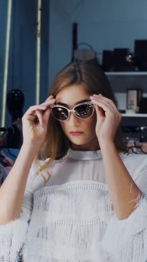 İş kadını şık bir mağazada güneş gözlüğü deniyor, butik yapıyor ve aynanın önünde kendine hayran, gülümsüyor. yavaş çekim.