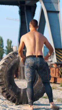Çıplak, çıplak gövdeli genç bir atletik, traktörden ağır bir lastik kaldırıyor, güç egzersizleri yapıyor, çapraz formda. Kumsalda, kargo limanında, büyük vinçlerin yanında, gün doğumu, yaz