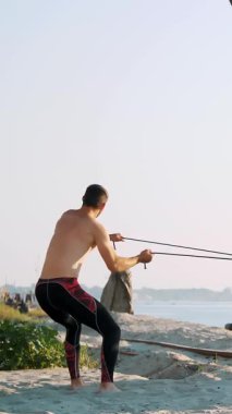 Çıplak, çıplak gövdeli genç bir atletik, kauçuk bir iple, çapraz bir elementle güç egzersizleri yapıyor. Kumsalda, bir kargo limanında, yazın, güneşin doğuşunda, büyük vinçlerin yanında.
