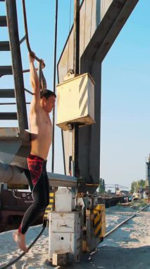 Çıplak gövdeli atletik bir adam, karın kasları için güç egzersizleri yapıyor, ayaklarını kaldırıyor, metal bir merdivende asılı duruyor. Bir kargo limanında, güneş doğarken, yazın.