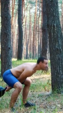 Atletik bir adam, çıplak, çıplak bir gövdeyle, ağırlık egzersizleri yapar. Çam ormanlarında, yazın, güneş ışınlarında