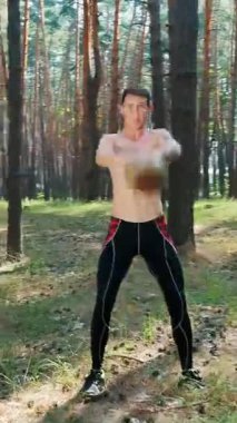 Atletik bir adam, çıplak, çıplak bir gövdeyle, ağırlık egzersizleri yapar. Çam ormanlarında, yazın, güneş ışınlarında