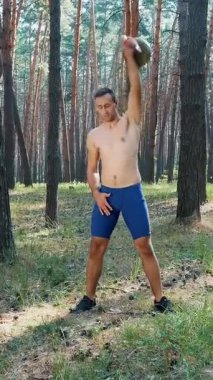 Atletik bir adam, çıplak, çıplak bir gövdeyle, ağırlık egzersizleri yapar. Çam ormanlarında, yazın, güneş ışınlarında