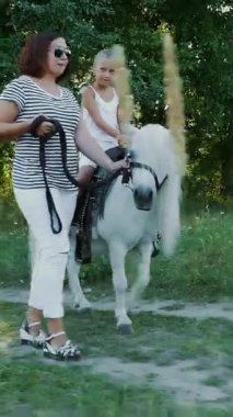 Kızı midilliye biniyor, annesi midilliyi dizginliyor. Neşeli, mutlu aile tatili. Açık havada, yazın, ormanın yakınında. Anne ve kızı tarlada yürüyorlar..