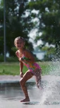 Mayo giymiş mutlu bir kız, çeşmedeki su sıçramaları arasında eğleniyor. Şehirde güneşli bir yaz günü. Yavaş çekim