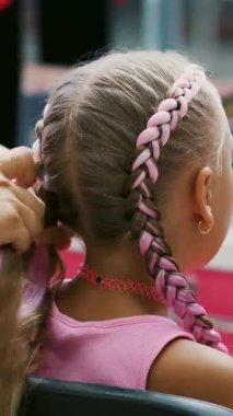 Yedi yaşında güzel sarışın bir kız, iki örgülü örgü örmüş, büyük bir aynanın önünde bir kuaför salonunda pembe bukleli saç stili yapmış. Küçük bir prenses.