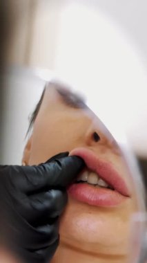 Kadın aynaya bakıyor, doktor dudaklarına hyaluronik asit enjekte ettikten sonra dudaklarının şeklini düzeltiyor. Sağlık Ofisi.