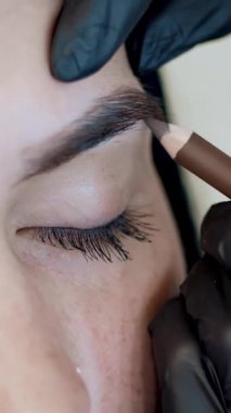 Kaş düzeltmesi. Yakın plan, kozmetik uzmanı, kaş yapıcı, siyah eldiven ustası, kaş boyası sürmeden önce kaş şeklini özel kalemle çiziyor. Kaşçının güzellik salonu..