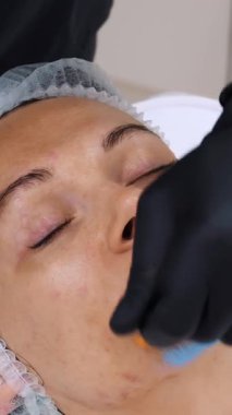 Yakın plan, siyah eldivenli kozmetik uzmanı, kadın yüzüne özel bir fırçayla mavi kozmetik yüz maskesi takıyor. Kozmetik kliniğinde veya güzellik salonunda cilt bakımı prosedürü.