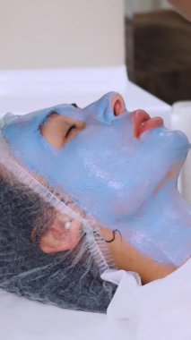 Yakın plan, siyah eldivenli kozmetik uzmanı, kadın yüzüne özel bir fırçayla mavi kozmetik yüz maskesi takıyor. Kozmetik kliniğinde veya güzellik salonunda cilt bakımı prosedürü.