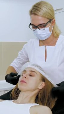 Kadınlar için kozmetik prosedürleri. Kozmetoloji. Kozmetik ameliyat için hazırlık. Güzellik uzmanı, estetik ameliyata başlamadan önce saçlarını saklayan bir kadının kafasına tek kullanımlık bir şapka takar. Güzellik kliniğinde. Cilt bakımı.