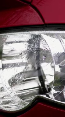 yakın, aracın far. duş, su sıçramalarına, kırmızı araba hood, cam far ıslak yüzeyi ile ağır damla düşer. Araba altında ıslak ağır yağmur alır,
