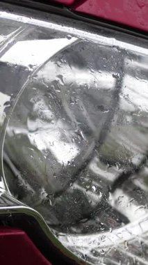 yakın, aracın far. duş, su sıçramalarına, kırmızı araba hood, cam far ıslak yüzeyi ile ağır damla düşer. Araba altında ıslak ağır yağmur alır,