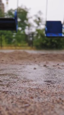 Çocuk parkına yağmur yağıyor. Yakın plan. Şiddetli yağmurda, yağmur damlaları su birikintilerine, su kabarcıklarına ve bahçeye düşer. Boş salıncak rüzgarda sallanıyor. Yağmur sırasında oyun alanını boşalt.