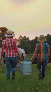 İnek sütü. İki çiftçi kadın, gün batımında otlayan ineklerin arka planına karşı yeşil çayır boyunca taze sütle dolu büyük bir konserve kutusu taşıyor. Süt sağmak. Süt çiftliği. Süt ürünleri. Çiftçilik.