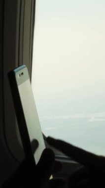Yakın plan. Çocuk eli ve cep telefonunun karanlık silueti uçağın aydınlatıcısına karşı. Çocuk kullanımı, akıllı telefonda oyun oynaması, uçak kabininde tablet telefon. Yolcular uçuş sırasında alet kullanıyorlar. Çocuklar için uçuş eğlencesi