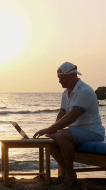 Spor şapkalı bir adam, dizüstü bilgisayar kullanıyor, gün batımında ya da gün doğumunda sahilde oturuyor. Serbest çalışan, uzaktan kumandalı. Denizde yaz tatili. İnternet mesafeleri siler