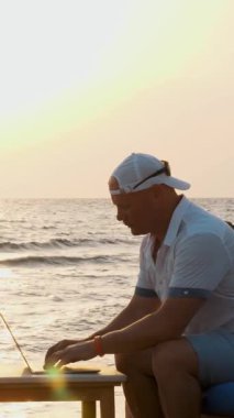 Spor şapkalı bir adam, dizüstü bilgisayar kullanıyor, gün batımında ya da gün doğumunda sahilde oturuyor. Serbest çalışan, uzaktan kumandalı. Denizde yaz tatili. İnternet mesafeleri siler